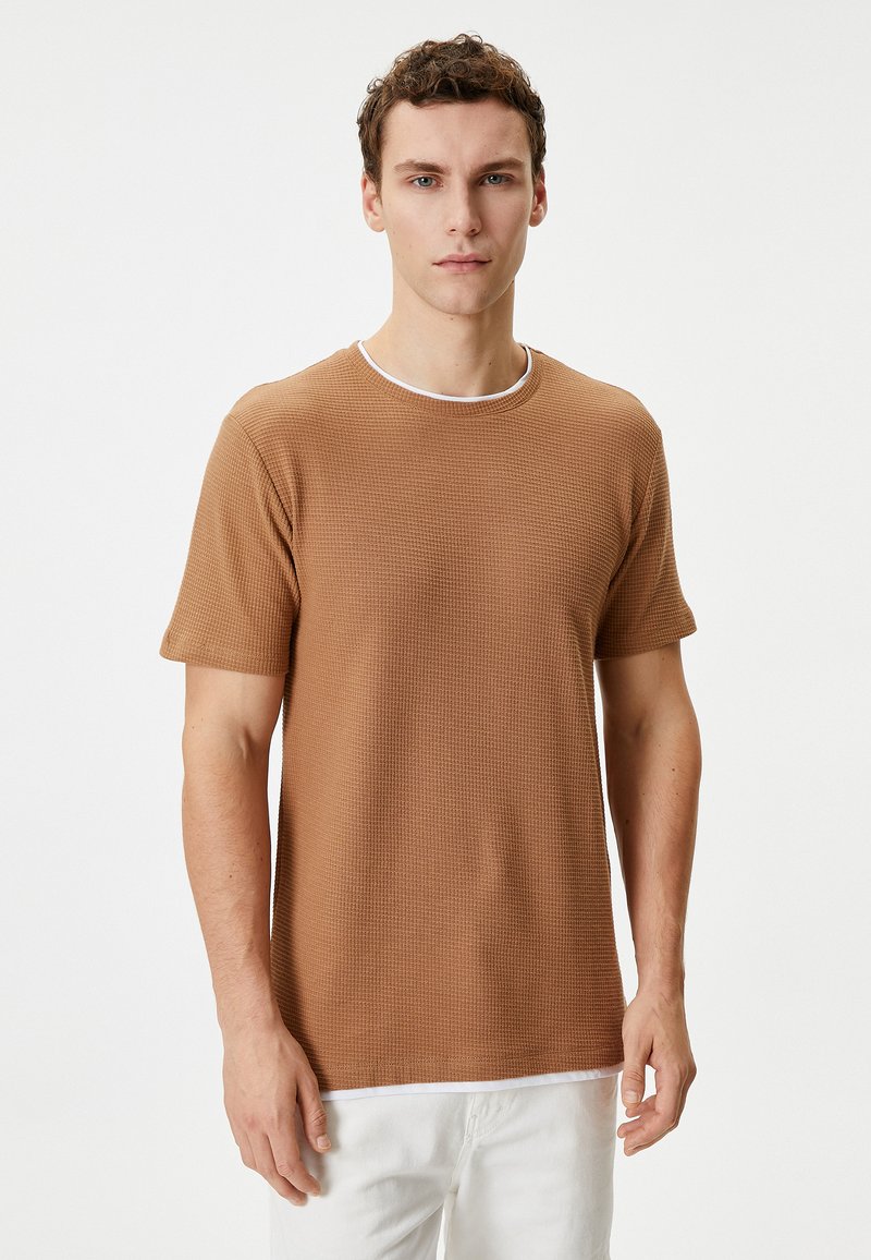 Brun tekstureret kortærmet t-shirt med rund halsudskæring, der har en hvid kant rundt om kraven, båret med lyse bukser.