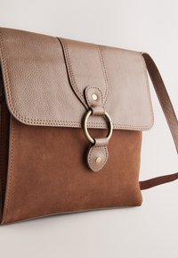 Sac bandoulière en cuir marron et daim avec fermeture à rabat et détail décoratif d'anneau en or sur le devant, sur fond uni.