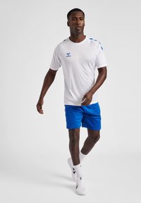 Witte sport t-shirt met blauwe accenten en logo, gecombineerd met blauwe shorts. Model draagt witte sneakers en sokken, terwijl hij/zij op een neutrale achtergrond loopt.