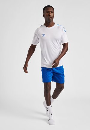 T-shirt de sport blanc avec des accents et un logo bleus, associé à un short bleu. Modèle portant des baskets blanches et des chaussettes, marchant sur un fond neutre.
