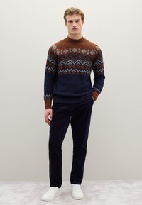 Gestrickter Pullover in Marineblau, Braun und Grau mit geometrischen Mustern, Rundhalsausschnitt und gerippten Bündchen, kombiniert mit dunkelblauen Cordhosen und weißen Turnschuhen.