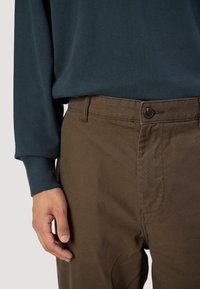 Brown pants with a button closure, made from structured fabric. The top is a dark teal long-sleeve shirt with cuffed sleeves.
