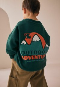 Grön sweatshirt med en grafisk design av berg och en helikopter i röd och vit, med texten "UTOMHUSÄVENTYR" i fet stil.