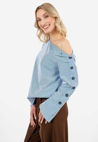 Blusa blu a righe, off-the-shoulder, con maniche ampie, caratterizzata da dettagli con bottoni lungo il braccio; abbinata a pantaloni marroni.