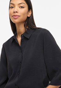 Naine pikkade tumedate juustega, kandes lahtist musta nööpidega särki, kerge naeratusega, ühevärvilise valge tausta ees.