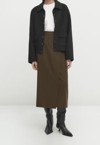 Schwarzwolljacke mit Druckknöpfen, getragen über einem weißen T-Shirt. Der olivgrüne Rock hat einen seitlichen Schlitz und wird mit schwarzen kniehohen Stiefeln mit Absatz kombiniert.