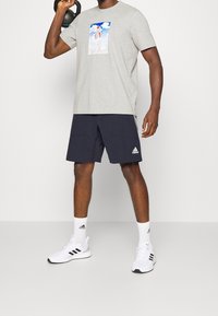 Grå t-shirt med kort ärm och grafiskt tryck, marinblå shorts och vita sportskor med svarta accenter. Figuren håller i en kettlebell.