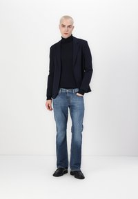 Navy blazer over a black turtleneck, paired with light blue flared jeans and black shoes. Smooth fabrics and tailored fit are noted.
