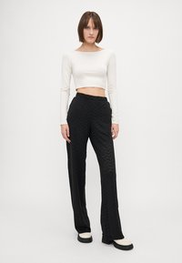 Černé kalhoty se zigzagovým vzorem a volným střihem, kombinované s upnutým bílým crop topem s dlouhým rukávem. Modelka má na sobě chunky boty s bílou podrážkou.