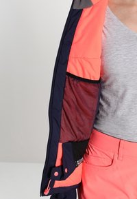 Osoba ubrana w koralowe spodnie i szarą koszulę trzyma otwartą granatową i koralową kurtkę, pokazując siatkową wewnętrzną kieszeń i zamek błyskawiczny.