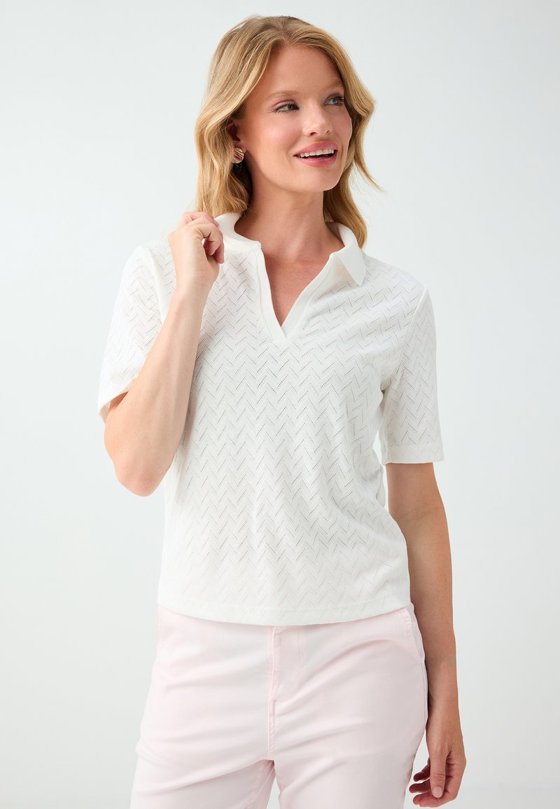 Femme blonde portant un polo blanc à manches courtes texturé et un pantalon rose pâle, souriante et regardant sur le côté sur un fond uni.