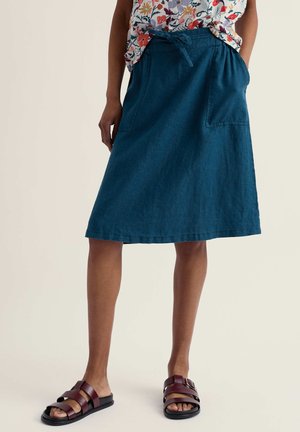 Persoană purtând o fustă albastră până la genunchi cu buzunare, un top floral introdus în fustă și sandale negre cu cataramă roșie, stând pe un fundal simplu.