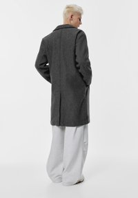 Szürke gyapjú kabát hosszú sziluettel, kivágott gallérral, lógó vállakkal és oldalsó zsebekkel, világosszürke, bő szárú nadrág felett viselve.