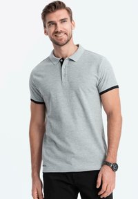 Světle šedé polo tričko s černým límcem a rukávy, tříknoflíkovou légou a decentním logem na lemu. Bavlněný materiál s hladkou texturou.