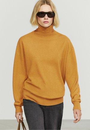 Kvinde iført sorte solbriller, en sennepsgul rullekravesweater, mørke jeans og med en brun skuldertaske mod en ensartet lys baggrund.