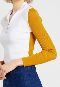 Ribbed long-sleeve top featuring a white body and mustard yellow sleeves with a half-zipper closure at the neck and fitted design.