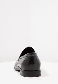 Mocassin noir en cuir sans lacets avec un petit talon et un détail subtil de couture bleue sur le talon, vu de l'arrière sur une surface et un fond blancs.