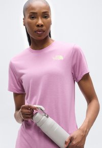 Γυναίκα που φοράει ροζ μπλουζάκι The North Face και κρατάει ένα ανοιχτό γκρι θερμομονωτικό μπουκάλι νερού με λαβή, σε απλό φόντο.