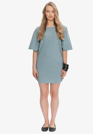 Młoda kobieta stoi przodem, ubrana w luźną, jasnoniebieską sukienkę z krótkimi rękawami, czarne bransoletki i czarne baleriny.