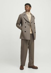 Manteau en laine gris à double boutonnage avec de grands boutons noirs, porté sur un pull en laine écru et un pantalon marron, accompagné de chaussures marron.