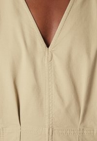 Gros plan sur un vêtement beige à col en V montrant des coutures verticales détaillées et la peau foncée du porteur au niveau de l'encolure.