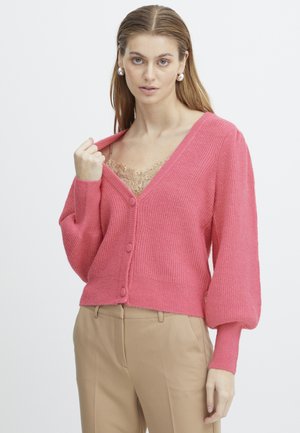 Gilet en tricot rose avec un décolleté en V, texture côtelée et manches ballons. Il est doté de boutons à l'avant et est superposé à un haut orné de dentelle.