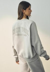 Világosszürke pulóver emelt "NEW YORK LEISURE CLUB" felirattal a hátsó részen, rakott ujjakkal és bordás mandzsettákkal, párosítva hozzá illő nadrággal.