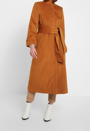 Femme portant un long manteau ceinturé couleur camel, un pantalon beige retroussé et des bottines blanches, debout devant un fond clair uni.