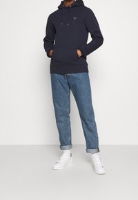 Uomo che indossa un hoodie blu navy con le mani nella tasca anteriore, jeans blu arrotolati, calze bianche e sneaker bianche, in piedi contro uno sfondo semplice.