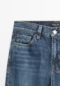 Donkerblauwe denim jeans met zichtbare stiksels, een knoopsluiting en een voorzak. Etiket zegt "Massimo Dutti, gemaakt in Turkije."