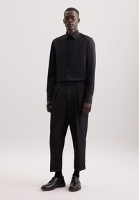 Camicia nera a maniche lunghe con bottoni abbinata a pantaloni a vita alta plissettati e accorciati. Le scarpe sono in pelle nera. Il materiale appare liscio e lucido.