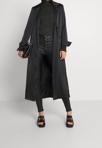 Długi czarny płaszcz satynowy o gładkiej fakturze, z zapięciem na guziki i szerokimi klapami. Nosi się go na błyszczącym czarnym golfie oraz czarnych spodniach z sztucznej skóry.