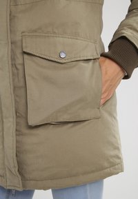 Beige bunda s velkou klopovou kapsou a patentním knoflíkem, kterou nosí osoba s rukou uvnitř boční kapsy, viditelný manžetový rukáv.