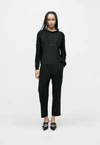 Felpa nera con cappuccio e coulisse regolabili, abbinata a pantaloni neri larghi e scarpe nere appuntite, su uno sfondo bianco.