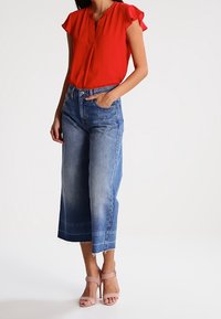 Blouse rouge à manches courtes et décolleté en V, associée à des culottes en denim bleu à jambes larges, avec un ourlet effiloché et un léger délavage, complétée par des sandales à talons roses.