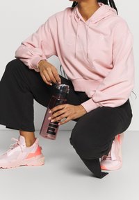 Sudadera rosa con un logo de Puma, pantalones negros y zapatillas rosa; sosteniendo una botella de agua transparente con texto negro. Ajuste relajado y casual.
