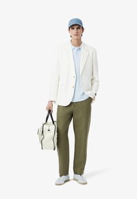 Witte blazer, lichtblauw polo-shirt, olijfgroene broek en witte sneakers. Houdt een crèmekleurige tas met zwarte accenten vast. Blauwe pet.