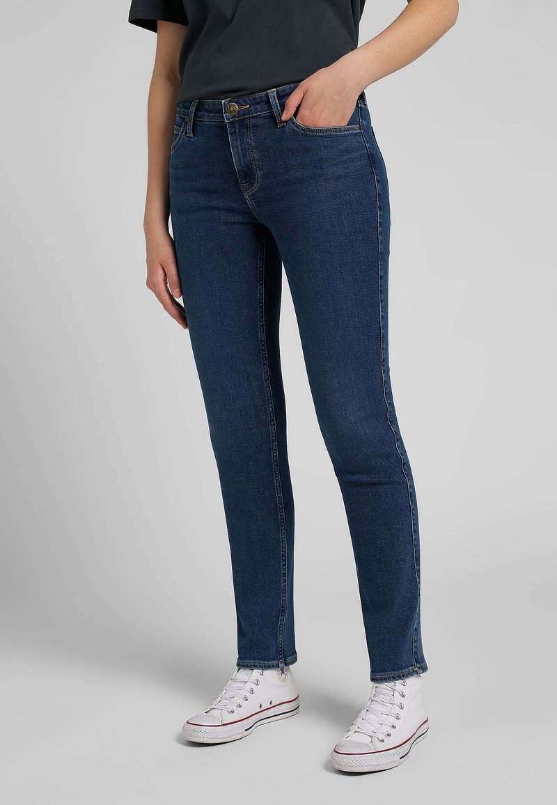 Dark blue denim skinny jeans with a smooth texture, five pockets, subtle stitching, and ankle-length hem paired with white sneakers.