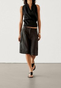 Schwarzes ärmelloses Wickeltop mit drapiertem Design, kombiniert mit lockeren, dunklen Shorts und flachen schwarzen Sandalen; minimalistischer Stil auf glattem Boden.