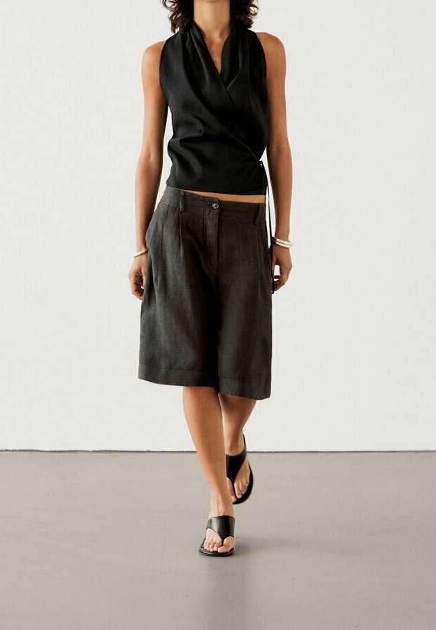 Schwarzes ärmelloses Wickeltop mit drapiertem Design, kombiniert mit lockeren, dunklen Shorts und flachen schwarzen Sandalen; minimalistischer Stil auf glattem Boden.