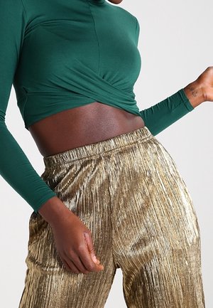 Osoba ubrana w zieloną, długą, krótką bluzkę z długim rękawem oraz wysoki stan błyszczących złotych plisowanych spodni, widziana od połowy tułowia do ud.