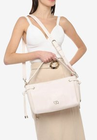 Witte leren handtas met verstelbare schouderriem, treksluiting en goudkleurige hardware. Kenmerkt zich door een subtiele reliëfdesign aan de voorkant.