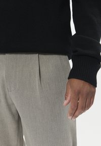 Melna adīta džemperis ar ribotiem aprocēm, pārī ar gaiši pelēkiem svītrainiem biksēm, kas raksturojas ar gludu tekstūru un sānu kabatām.