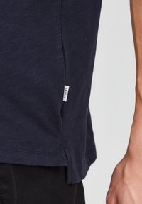 Tricou din bumbac de culoare bleu marin, cu fente laterale, având un eticheta țesută inscripționată cu "VINERI" pe partea inferioară. Textură netedă și croi lejer.