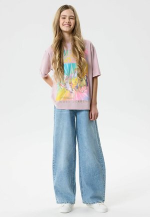 Ragazza sorridente con lunghi capelli biondi che indossa una t-shirt grafica con unicorno pastello, jeans a gamba larga azzurri chiaro e sneakers bianche, in piedi con le mani dietro la schiena.