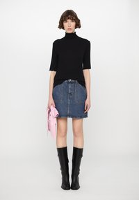 Malha preta com gola alta, mangas curtas, combinada com uma saia A-line de denim. Botas pretas até ao joelho e uma camisa rosa pendurada no braço.