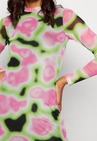 Gros plan sur une personne portant une robe ajustée à manches longues avec un motif tie-dye flou dans des couleurs rose, vert, noir et blanc.