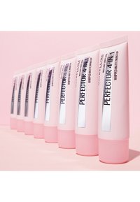 Dieci tubi rosa del fondotinta mattificante "Perfector 4-in-1" di Maybelline sono rivolti verso l'avanti, ognuno con un'etichetta trasparente e accenti metallici, su uno sfondo rosa.