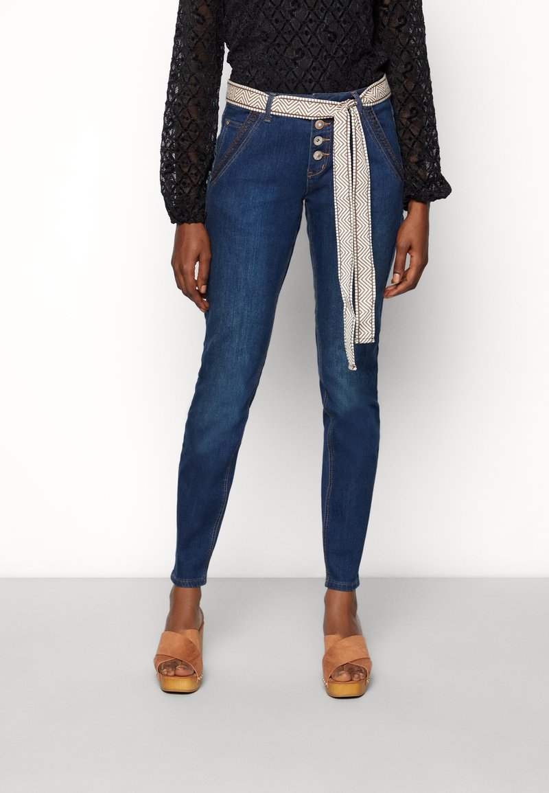 Jean skinny en denim bleu foncé avec une ceinture beige à motifs, des boutons en détail, et un haut noir texturé. Des sandales à talons marron complètent le look.