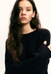 Fekete kötött pulóver kerek nyakkivágással és bordázott mandzsettákkal, puha textúrával. Hosszú, hullámos haj részben takarja a vállat.